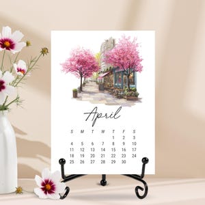Puede incluir: Una p&aacute;gina de calendario vertical para abril con una ilustraci&oacute;n en acuarela de una escena callejera con cerezos en flor de color rosa, asientos de caf&eacute; al aire libre y un escaparate. La cuadr&iacute;cula del calendario muestra los d&iacute;as del mes.