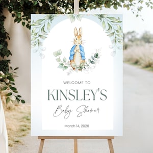 May include: A white welcome sign on an easel, featuring a watercolor illustration of Peter Rabbit in a blue jacket. The sign reads "WELCOME TO KINSLEY'S Baby Shower" with the date March 14, 2026.