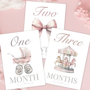 May include: Three pink and white baby milestone cards. Each card features a watercolor illustration and text. The cards display the words "One Month", "Two", and "Three Months". The illustrations include a baby carriage, a bow, and a carousel.