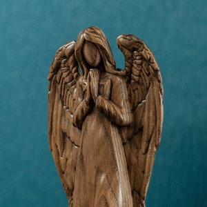 May include: A wooden angel figurine with wings outstretched and hands clasped in prayer. The angel is carved from a light brown wood and has a detailed, realistic appearance.