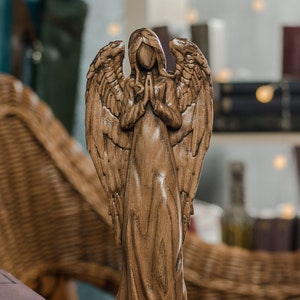 Wooden angel statue with folded hands standing on a wooden table, carved from light natural wood, with detailed wings and soft window light creating a peaceful, spiritual atmosphere