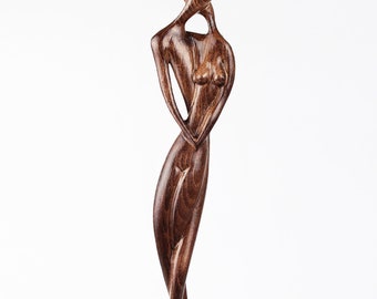 May include: A wooden sculpture of two figures embracing, carved in a dark brown color. The sculpture is abstract and stylized, with the figures' bodies intertwined. The sculpture is standing on a wooden base.