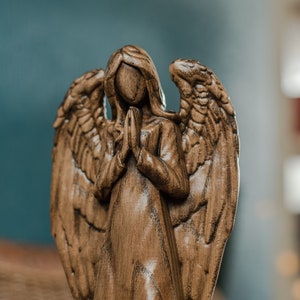 May include: A wooden angel figurine with wings outstretched and hands clasped in prayer. The angel is carved from dark brown wood and has a detailed, realistic appearance.