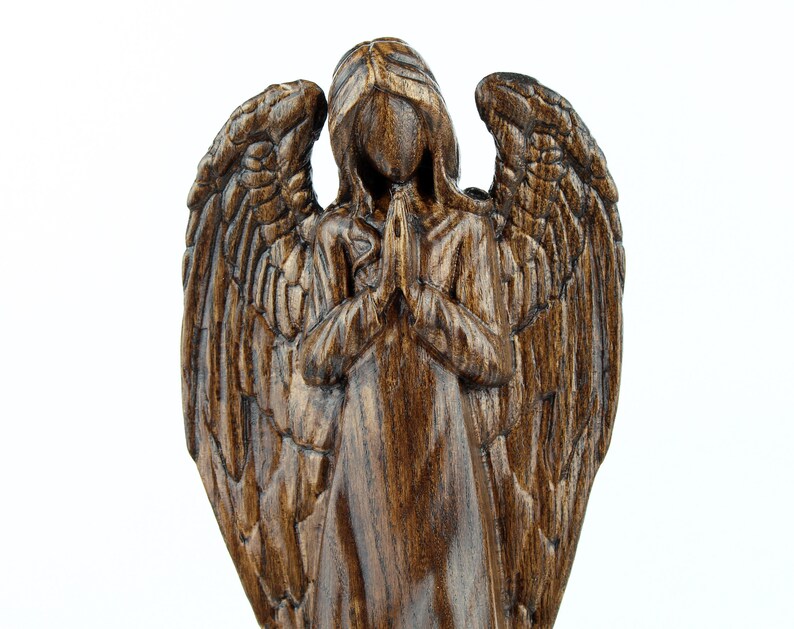 May include: A dark brown wooden angel figurine with wings outstretched and hands clasped in prayer. The angel is carved in a realistic style with detailed features.