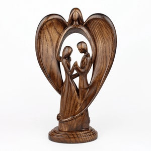 May include: A wooden sculpture of a couple standing inside a heart-shaped frame with wings. The sculpture is made of dark brown wood and has a rustic finish.