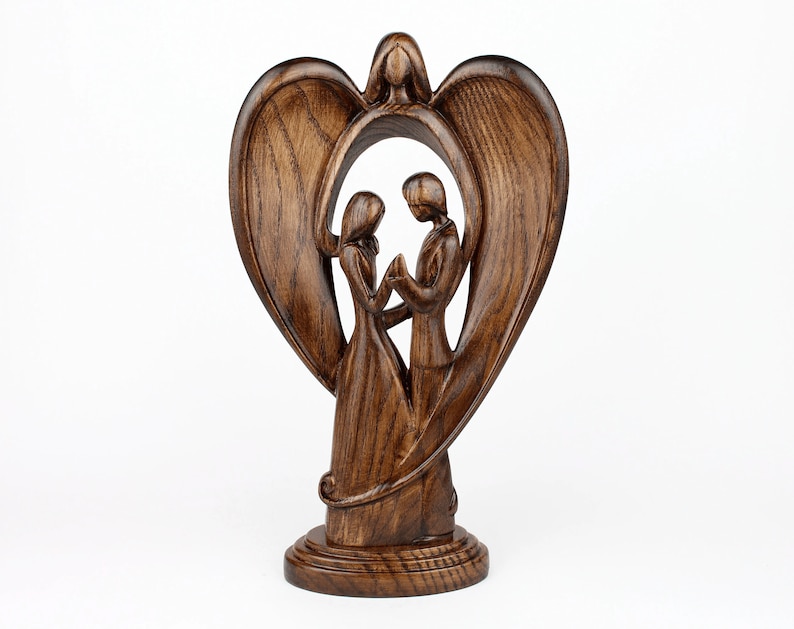 May include: A wooden sculpture of a couple standing inside a heart-shaped frame with wings. The sculpture is made of dark brown wood and has a rustic finish.