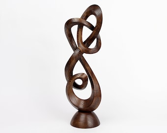 Puede incluir: Una escultura de madera marrón oscuro con un diseño abstracto en espiral. La escultura está sobre una base redonda y es alta.
