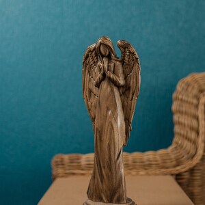 May include: A wooden angel figurine with wings outstretched and hands clasped in prayer. The angel is standing on a wooden base and is carved with intricate details. The figurine is brown in color and has a rustic finish.