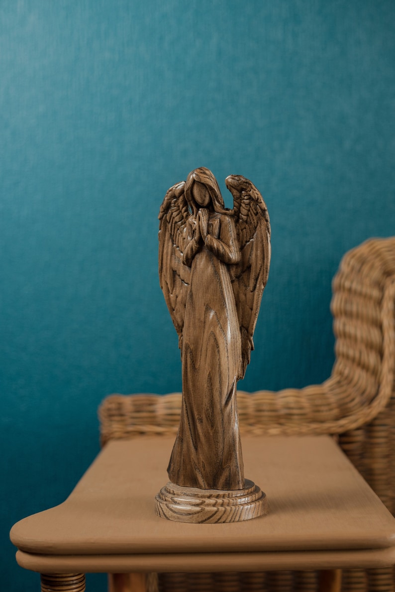 May include: A wooden angel figurine with wings outstretched and hands clasped in prayer. The angel is standing on a wooden base and is carved with intricate details. The figurine is brown in color and has a rustic finish.