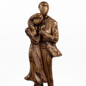 May include: A wooden sculpture of a couple embracing. The figures are carved from a light brown wood, with visible grain patterns. The man is taller, and the woman is leaning into him. The sculpture is on a white background.