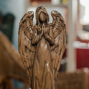 May include: A wooden angel figurine with outstretched wings, carved in a detailed style. The angel is standing with hands clasped in prayer, and the wood has a rich brown color.