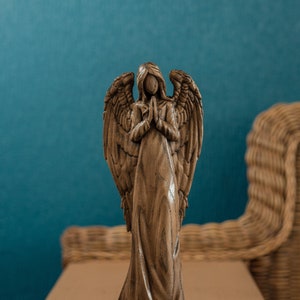 May include: A wooden angel figurine with outstretched wings, standing on a wooden base. The angel is carved in a prayer pose with hands clasped together. The figurine is painted in a dark brown color.