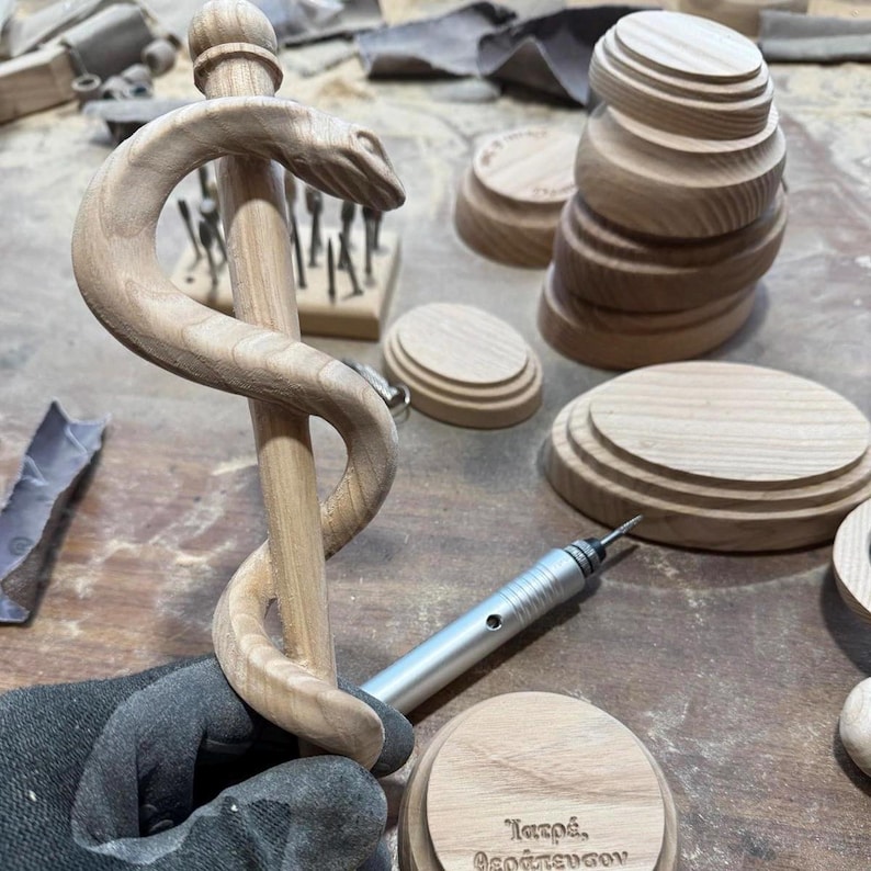 May include: A wooden medical symbol, the Rod of Asclepius, featuring a serpent coiled around a staff. The image shows the symbol in progress, with other wooden components and tools nearby. The wood has a natural, light brown color.