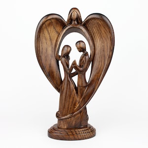 May include: A wooden sculpture of a couple standing inside a heart-shaped frame with wings. The sculpture is made of dark brown wood and has a rustic finish.