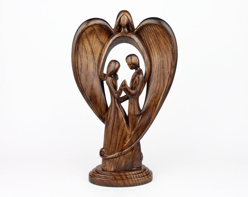 May include: A wooden sculpture of a couple standing inside a heart-shaped frame with wings. The sculpture is made of dark brown wood and has a rustic finish.