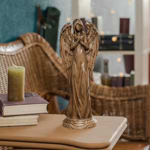 May include: A wooden angel figurine with outstretched wings, standing on a wooden base. The angel is carved in a realistic style and has a serene expression. The figurine is placed on a wooden table with a candle and books.