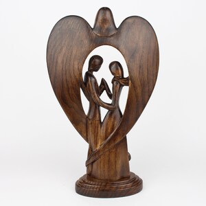 May include: A wooden sculpture of a couple standing inside a heart-shaped frame. The sculpture is made of dark brown wood and has a smooth finish.