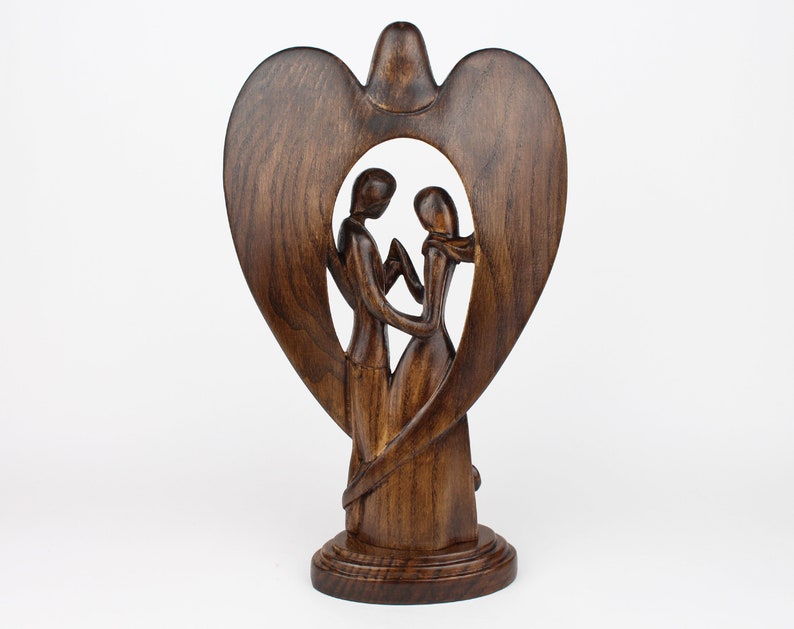 May include: A wooden sculpture of a couple standing inside a heart-shaped frame. The sculpture is made of dark brown wood and has a smooth finish.