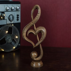 May include: A wooden sculpture of a treble clef with a heart shape carved into the center. The sculpture is brown with a wood grain pattern and is standing on a wooden base.