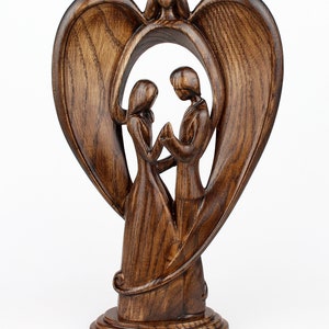 May include: A wooden sculpture of a couple standing inside a heart-shaped frame with angel wings. The sculpture is made of dark brown wood and has a rustic finish.