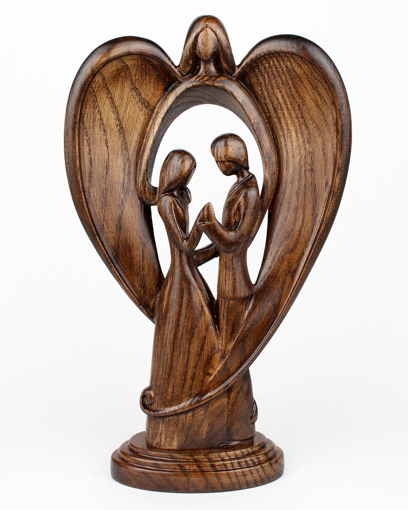 May include: A wooden sculpture of a couple standing inside a heart-shaped frame with angel wings. The sculpture is made of dark brown wood and has a rustic finish.