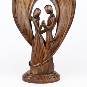 May include: A wooden sculpture of a couple standing inside a heart-shaped frame. The sculpture is made of dark brown wood and has a smooth, polished finish.
