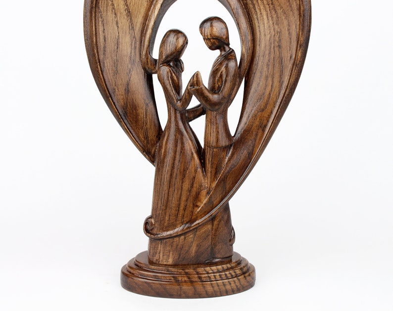 May include: A wooden sculpture of a couple standing inside a heart-shaped frame. The sculpture is made of dark brown wood and has a smooth, polished finish.