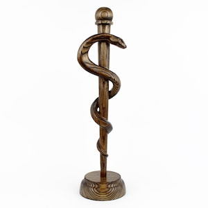 Op de afbeelding: Een houten caduceus sculptuur, met een staf met een slang eromheen gewikkeld. De sculptuur is gemaakt van donkerbruin hout en heeft een ronde basis en een bal aan de bovenkant. De kop van de slang is zichtbaar.