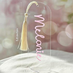 Puede incluir: Un marcapáginas de acrílico transparente con bordes redondeados, que presenta el nombre Melanie en letras rosas. Una borla de color crema está unida al orificio superior. El marcapáginas mide 2 pulgadas de ancho, 5,8 pulgadas de alto y 1,5 mm de grosor.