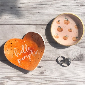 Puede incluir: Dos cuencos de cerámica para baratijas. Uno tiene forma de corazón con un degradado naranja y la frase "hello pumpkin". El otro es redondo con interior rosa e ilustraciones de calabazas. Un par de anillos están cerca.
