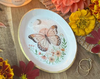 May include: An oval white ceramic trinket dish with a gold-painted rim, featuring a central illustration of a butterfly, a crescent moon, and small flowers. Two thin gold rings rest beside the dish on a wooden surface with scattered flower petals.