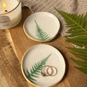 May include: Two round ceramic trinket dishes with a white speckled glaze and a hand-painted green fern leaf design. Each dish features a textured gold-painted rim. One dish contains two gold rings resting on its surface.