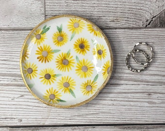 Puede incluir: Un pequeño plato de cerámica blanca con un borde dorado y un patrón de girasoles amarillos. El plato está sobre una superficie de madera blanca.