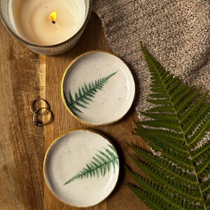 May include: Two round ceramic trinket dishes with a speckled white glaze and a green fern leaf print. Each dish features a metallic gold painted rim. The dishes are arranged alongside two gold rings, a lit candle, and a green fern frond.