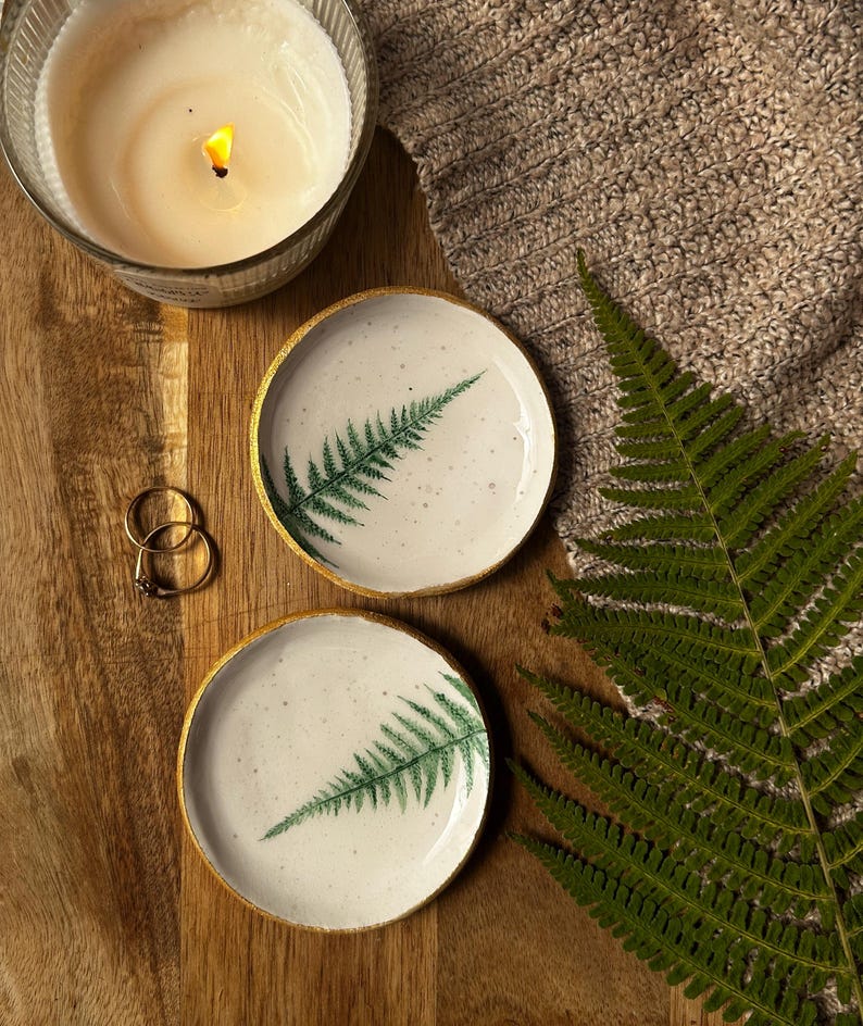May include: Two round ceramic trinket dishes with a speckled white glaze and a green fern leaf print. Each dish features a metallic gold painted rim. The dishes are arranged alongside two gold rings, a lit candle, and a green fern frond.
