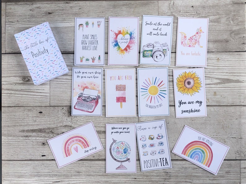 Puede incluir: Una caja de tarjetas de afirmaci&oacute;n de positividad con ilustraciones coloridas y mensajes inspiradores. Las tarjetas presentan una variedad de dise&ntilde;os, incluyendo arco&iacute;ris, girasoles y una m&aacute;quina de escribir. La caja es blanca con un patr&oacute;n de confeti colorido y las palabras "the little box of positivity" impresas en ella.
