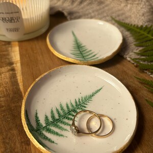 May include: Two round white ceramic trinket dishes featuring a hand-painted green fern leaf design and gold-painted edges. One dish holds two gold rings. The surface of the dishes has a speckled texture, and the edges are uneven and organic.