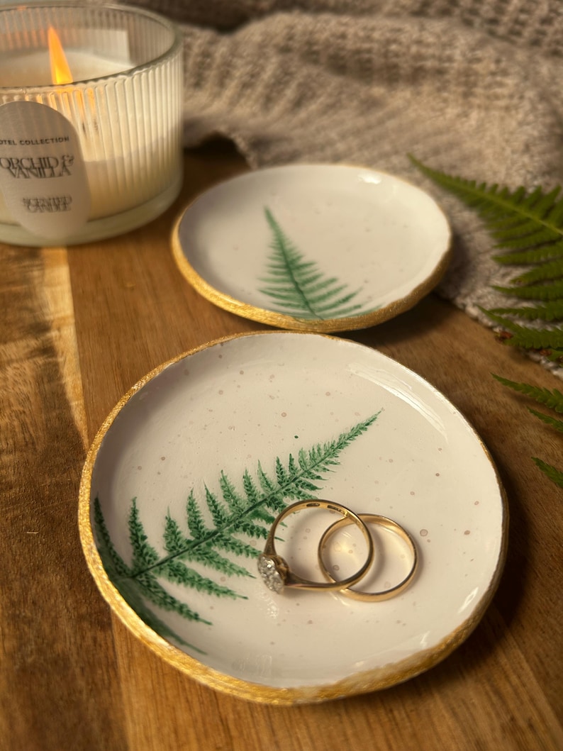 May include: Two round white ceramic trinket dishes featuring a hand-painted green fern leaf design and gold-painted edges. One dish holds two gold rings. The surface of the dishes has a speckled texture, and the edges are uneven and organic.
