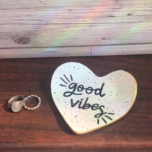 May include: White heart-shaped ceramic dish with gold speckles and black lettering that reads "good vibes".