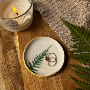 May include: A small, round, white ceramic trinket dish featuring a green fern leaf illustration and a metallic gold painted rim. Two thin gold rings rest inside the dish. The surface is speckled with tiny dark dots, and the edges are slightly uneven.