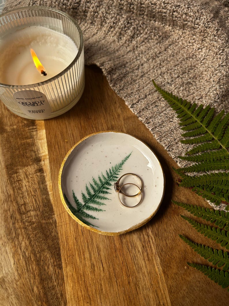 May include: A small, round, white ceramic trinket dish featuring a green fern leaf illustration and a metallic gold painted rim. Two thin gold rings rest inside the dish. The surface is speckled with tiny dark dots, and the edges are slightly uneven.