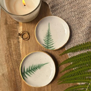 May include: Two round ceramic trinket dishes with a white speckled glaze and a green fern leaf print. Each dish features a metallic gold painted rim. The dishes are displayed alongside a lit candle, two gold rings, and a green fern frond.