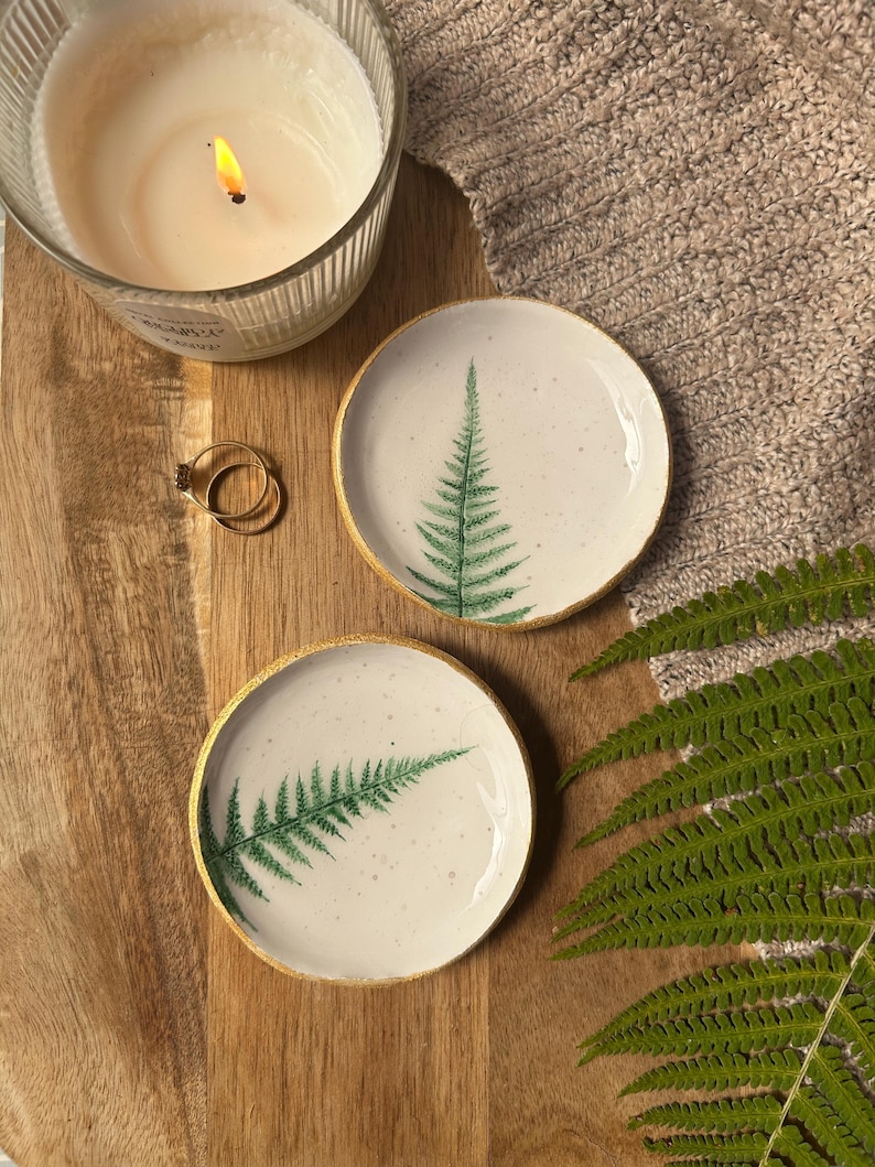 May include: Two round ceramic trinket dishes with a white speckled glaze and a green fern leaf print. Each dish features a metallic gold painted rim. The dishes are displayed alongside a lit candle, two gold rings, and a green fern frond.