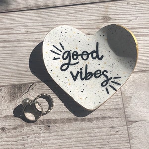 May include: White heart-shaped ceramic dish with black speckles and gold trim. The dish has the words "good vibes" written in black cursive lettering.