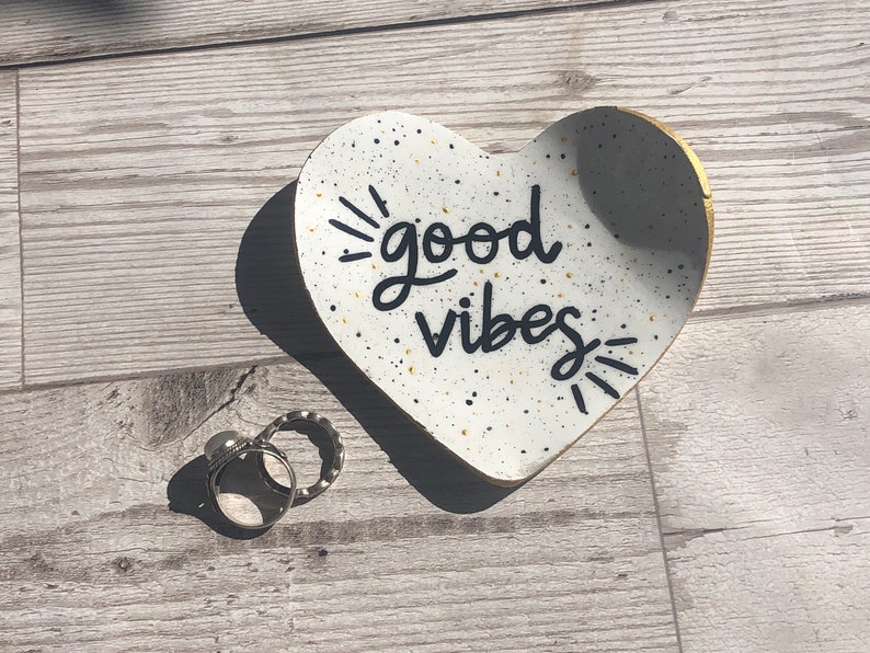 May include: White heart-shaped ceramic dish with black speckles and gold trim. The dish has the words "good vibes" written in black cursive lettering.