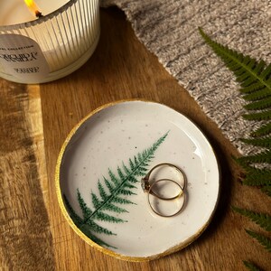 May include: A round white ceramic trinket dish featuring a green fern leaf illustration and a gold-painted rim. Two thin gold rings rest inside the dish. The surface of the dish has small dark speckles throughout its white glazed finish.