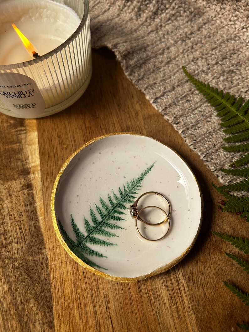 May include: A round white ceramic trinket dish featuring a green fern leaf illustration and a gold-painted rim. Two thin gold rings rest inside the dish. The surface of the dish has small dark speckles throughout its white glazed finish.