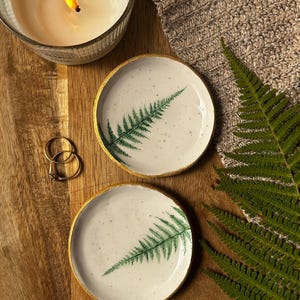 May include: Two round ceramic trinket dishes with a speckled white surface and a green fern leaf print. Each dish features a metallic gold painted rim. The dishes are circular with a smooth, shallow basin design, showcasing a botanical aesthetic.