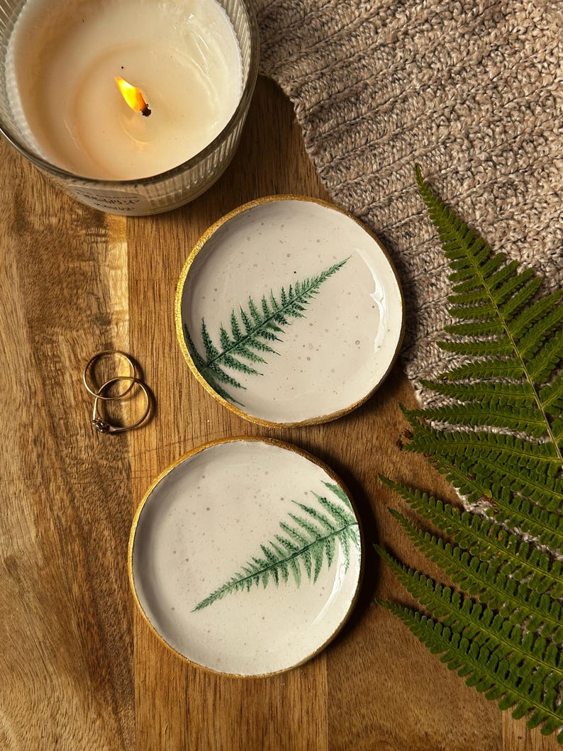 May include: Two round ceramic trinket dishes with a speckled white surface and a green fern leaf print. Each dish features a metallic gold painted rim. The dishes are circular with a smooth, shallow basin design, showcasing a botanical aesthetic.