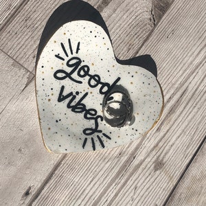 May include: White heart-shaped ceramic dish with black speckles and gold glitter. The dish has the words "Good Vibes" written in black cursive. There are several silver rings in the dish.